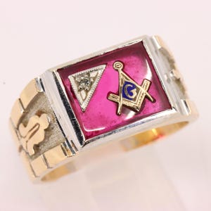 May include: A two-tone gold and silver ring featuring a rectangular face with a deep pink inlay. The face displays a small diamond-set triangle and a gold Masonic square and compass emblem with a blue letter G in the center of the design.