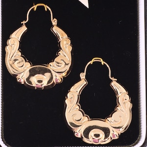 May include: A pair of gold-colored hoop earrings. Each earring features an ornate, detailed design with a central oval shape and curved, decorative elements. The earrings are displayed against a black background.