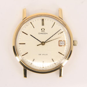 May include: A gold Omega De Ville wristwatch with a cream dial. The watch has black hands and markers, a date window at 3 o'clock, and the Omega logo. The round watch case has lugs for a strap. The watch is a classic design.