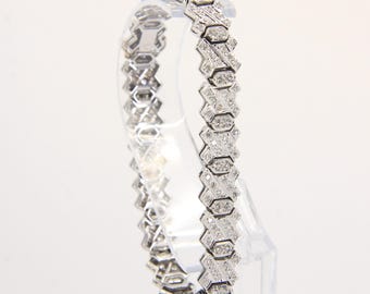 Könnte beinhalten: Ein silbernes Armband mit geometrischem Design. Das Armband besteht aus miteinander verbundenen, X-förmigen Gliedern, die jeweils mit kleinen, funkelnden Edelsteinen besetzt sind. Das Armband wird auf einem transparenten Ständer vor weißem Hintergrund präsentiert.