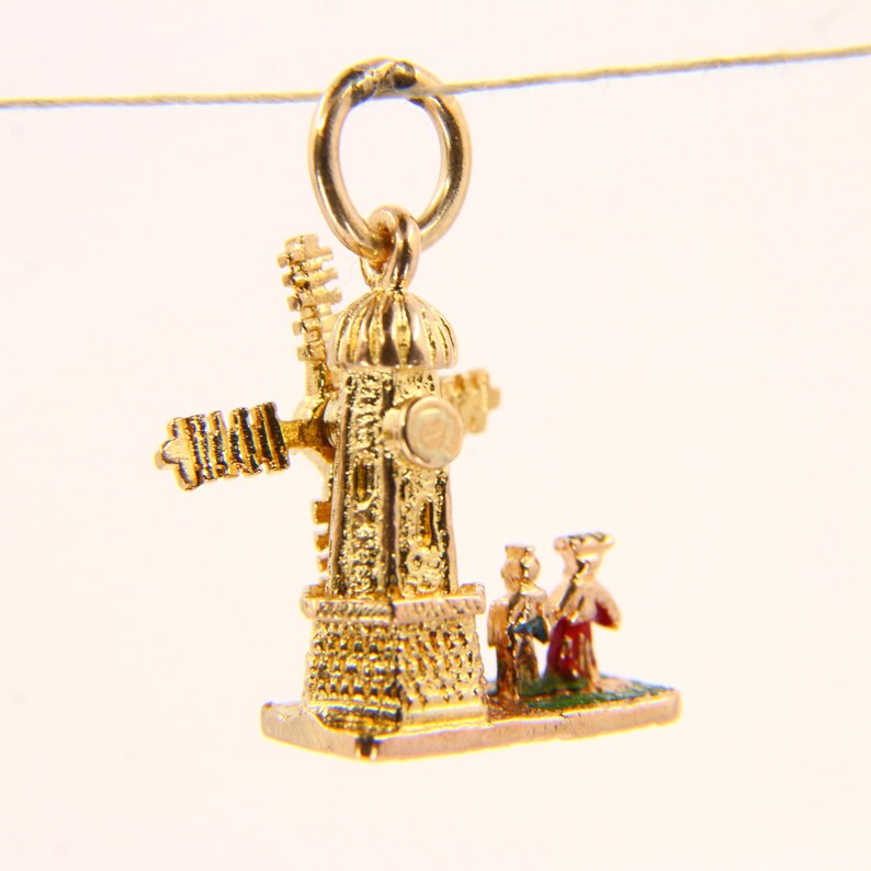 May include: A gold charm in the shape of a windmill with two figures standing in front of it. The windmill has a gold roof and a gold base. The figures are wearing red and blue clothing.