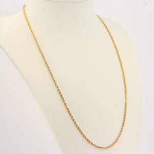 Puede incluir: Un collar de cadena de cuerda de color dorado exhibido en un maniquí blanco. La cadena está hecha de pequeños eslabones tejidos, creando una apariencia texturizada. El collar está drapeado en un estilo largo y elegante.