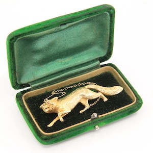 Puede incluir: Broche de zorro dorado con cadena, exhibido en una caja de joyería vintage de terciopelo verde. El zorro está detallado con pelaje texturizado y una pose de carrera. La caja tiene una tapa con bisagras y un interior verde oscuro.
