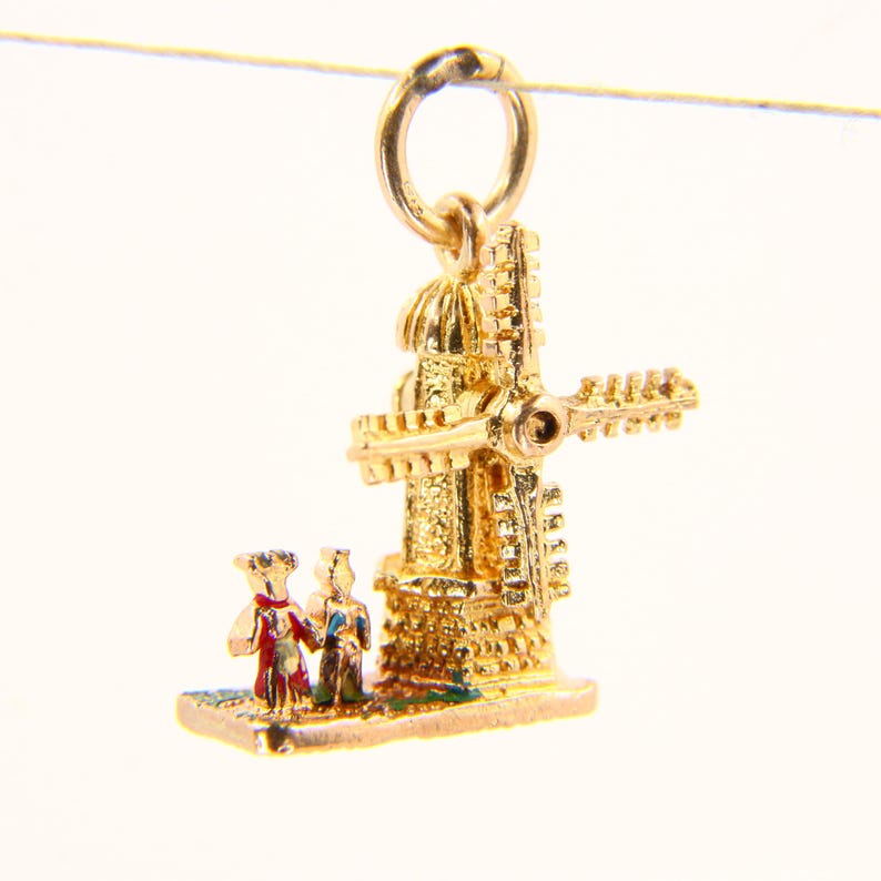 May include: Gold charm of a windmill with a couple standing in front of it. The windmill is detailed with gold and enamel accents.