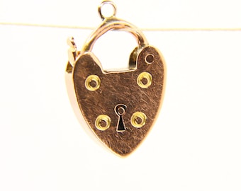 May include: A heart-shaped gold-colored padlock charm with a keyhole and four decorative rivets. The charm has a small loop at the top for hanging. The padlock is suspended against a white background.
