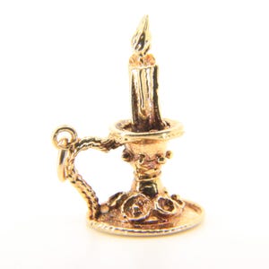 Puede incluir: Un dije dorado con forma de vela con un dise&ntilde;o detallado. El dije presenta una vela con una llama, colocada en un candelabro decorativo con un asa. El dije probablemente est&aacute; hecho de oro o metal dorado.