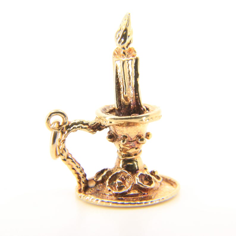 Puede incluir: Un dije dorado con forma de vela con un dise&ntilde;o detallado. El dije presenta una vela con una llama, colocada en un candelabro decorativo con un asa. El dije probablemente est&aacute; hecho de oro o metal dorado.