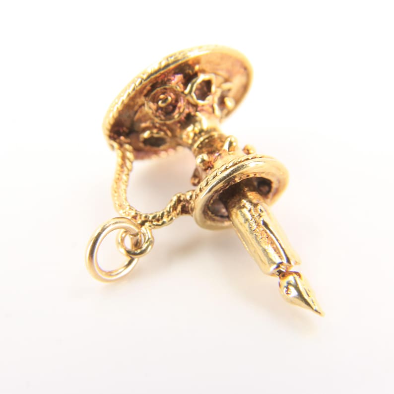 Puede incluir: Un dije dorado en forma de candelabro con una pluma. El dije tiene un dise&ntilde;o detallado, que incluye un asa en forma de cuerda y elementos decorativos en la parte superior e inferior. La pluma tiene una punta.