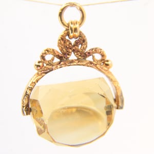 Puede incluir: Un colgante dorado con una gran gema amarilla facetada. El colgante tiene un diseño ornamental de estilo filigrana alrededor de la gema y un lazo en la parte superior para una cadena. La gema tiene aproximadamente 2,5 cm de diámetro.