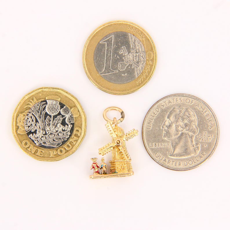 May include: Gold charm of a windmill with two figures standing in front of it. The charm is about the size of a US quarter.