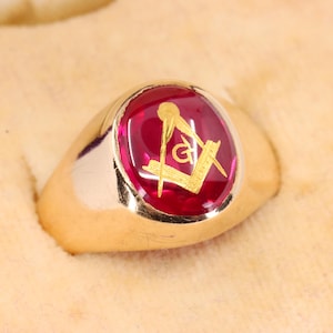 May include: A gold ring featuring a smooth, oval-shaped deep red stone set in the center. The surface of the red stone displays a gold-colored Masonic square and compass emblem. The band of the ring is polished gold with a solid, rounded design.
