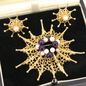 Op de afbeelding: Een goudkleurige sieradenset met een grote broche en twee bijpassende oorbellen. De broche is versierd met paarse en witte kralen, terwijl de oorbellen en de broche een sterrenpatroon hebben. De set wordt gepresenteerd in een zwarte sieradendoos.