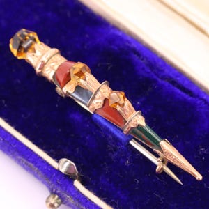 May include: A decorative brooch in the shape of a dagger, featuring gold-colored metalwork, amber-colored gemstones, and sections of red, green, and black enamel. The brooch is presented on a blue velvet surface.