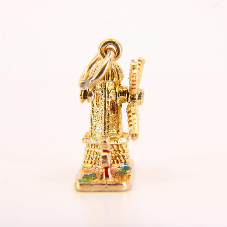May include: A gold charm in the shape of a windmill with a red roof and green and yellow accents. The charm is detailed with a small windmill blade and a loop for attaching to a necklace or bracelet.