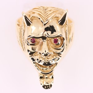 May include: A gold-colored ring featuring a devil face design. The devil face has horns, a detailed facial structure, and two red gemstones for eyes. The ring is likely made of gold and has a polished finish.
