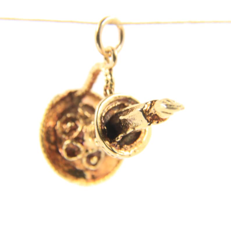 Puede incluir: Un dije dorado que representa una sart&eacute;n en miniatura con un asa y un peque&ntilde;o utensilio adjunto. El dije est&aacute; suspendido de un peque&ntilde;o lazo, lo que sugiere que est&aacute; dise&ntilde;ado para un collar o pulsera. El dise&ntilde;o es intrincado.
