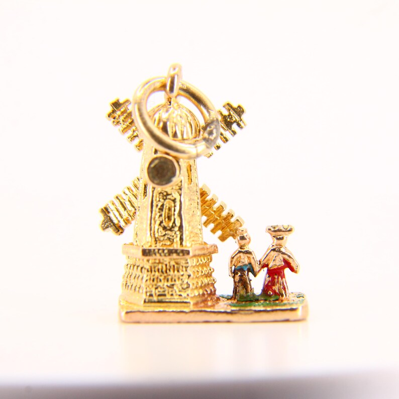 May include: Gold charm of a windmill with a red roof and two figures in front of it. The figures are wearing colorful clothing and are holding hands.