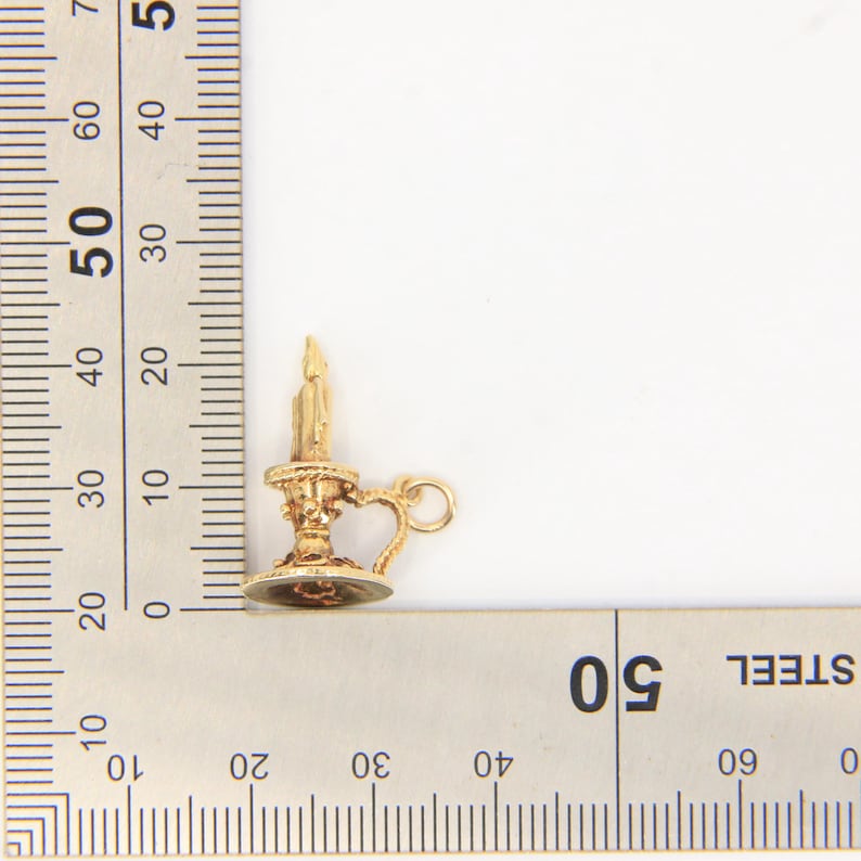 Puede incluir: Un candelabro en miniatura de color dorado con un asa y un peque&ntilde;o lazo. La vela est&aacute; encendida y el soporte tiene detalles intrincados. El art&iacute;culo se muestra junto a una regla para la escala.