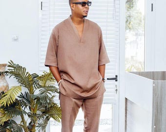 May include: A brown linen two-piece suit with a V-neck top and matching trousers. The outfit is worn with black sandals and sunglasses. A potted plant and a window with blinds are in the background.