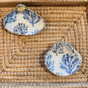 Puede incluir: Dos platos con forma de concha con fondo blanco y diseños de coral azul. Los bordes están adornados con dorado. Los platos se exhiben en una bandeja de mimbre tejido.