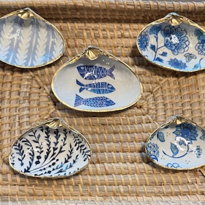 Puede incluir: Cinco conchas decorativas con bordes dorados. Los diseños interiores varían, con patrones azules y blancos que incluyen peces, motivos florales y elementos botánicos. Las conchas están dispuestas sobre una bandeja tejida.