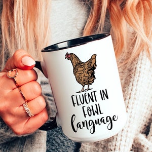 Puede incluir: Taza de cerámica blanca con borde y asa negros. La taza presenta una ilustración de pollo marrón y el texto "Fluent in fowl language".