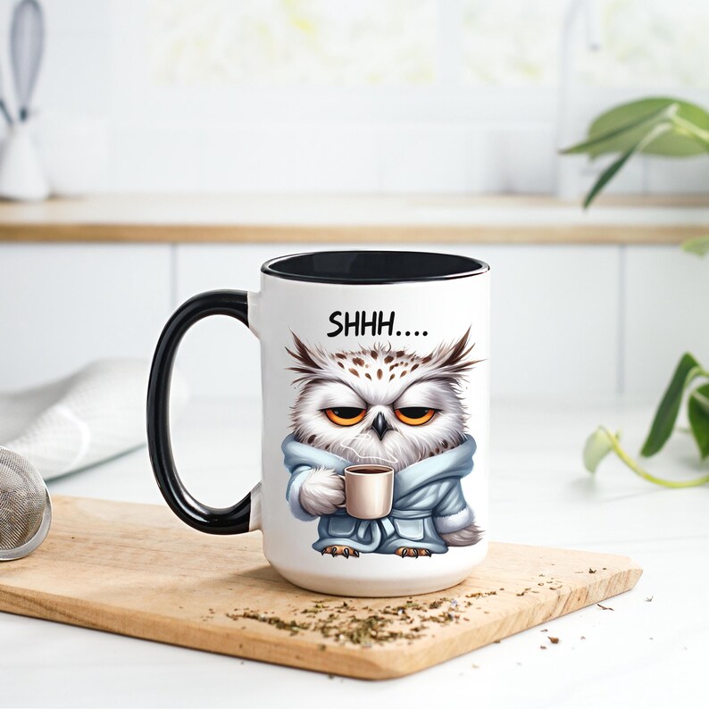 May include: White ceramic mug with a black handle and rim, featuring an illustration of an owl in a blue robe holding a coffee cup. The mug has the text "SHHH...." printed on it. The mug is sitting on a wooden cutting board.