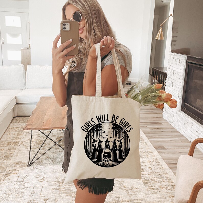 May include: A white canvas tote bag with black text that reads "Girls Will Be Girls" and a black and white illustration of witches in a forest.
