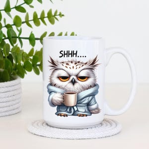 May include: White ceramic mug featuring a grumpy owl wearing a blue robe and holding a coffee cup. The mug has the text "SHHH...." above the owl. The mug is on a white coaster.