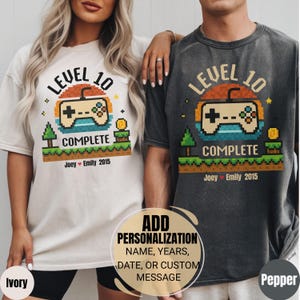 May include: Two t-shirts, one ivory and one pepper-coloured, featuring a pixelated video game controller design. The text reads "LEVEL 10 COMPLETE" with "Joey Emily 2015" below. Personalisation options are available for the shirts.