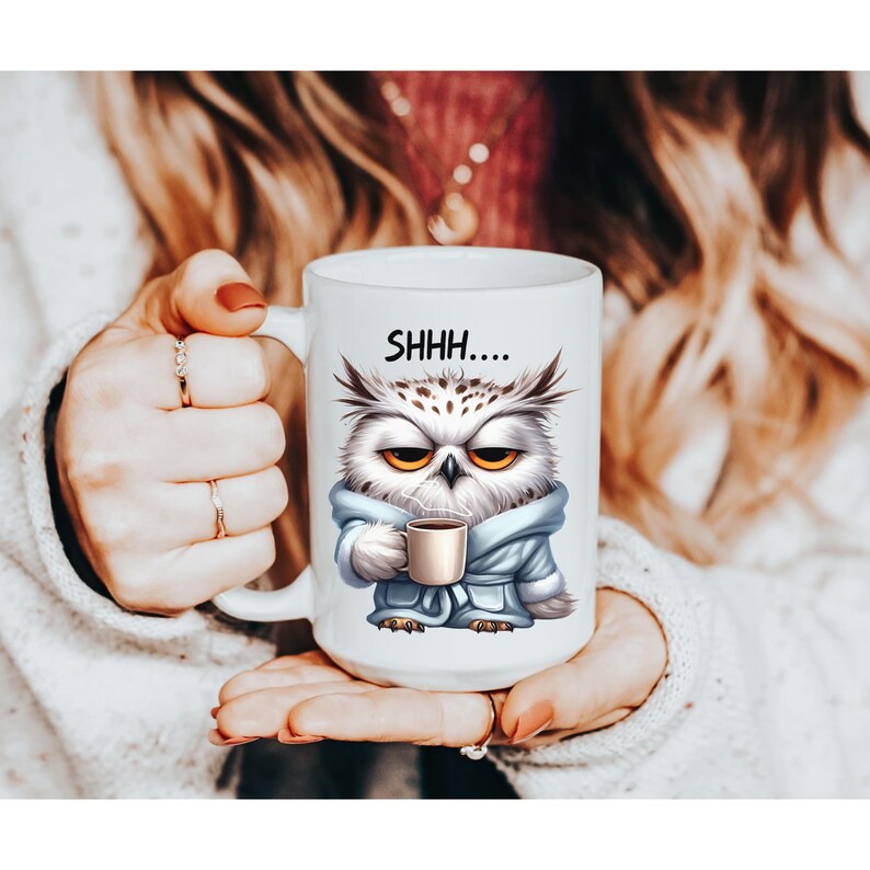 May include: White ceramic mug featuring a cartoon owl in a blue robe, holding a coffee cup. The owl has yellow eyes and the text "SHHH...." is printed on the mug. The mug is held by a person.