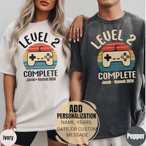 Puede incluir: Dos camisetas, una marfil y otra pimienta, con un diseño de juego retro. El diseño incluye el texto "LEVEL 2 COMPLETE" con un gráfico de mando y los nombres "Jacob Hannah 2024". Las camisetas son para parejas.