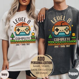 May include: Ivory and pepper-colored t-shirts with a pixelated video game controller design. The shirts read "LEVEL 8 COMPLETE" with the names "Joey Emily 2017" below. The design includes a pixelated sunset and trees. The shirts are customizable.