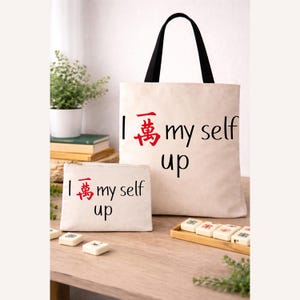 May include: An off-white canvas tote bag and a matching zippered pouch, both printed with the phrase "I 萬 my self up" in black and red. The tote bag has black handles. The items are displayed on a wooden surface.