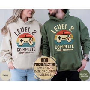 May include: Two matching hoodies, one sand-colored and one military green, feature a retro video game controller design. The text reads "LEVEL 2 COMPLETE" with a personalized name and year. The design is set against a sunset-colored circle.