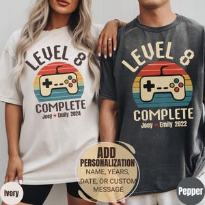 May include: Ivory and pepper-colored t-shirts with a retro video game controller design. The shirts feature the text "LEVEL 8 COMPLETE" with personalized names and dates. The design includes a sunset-style background.