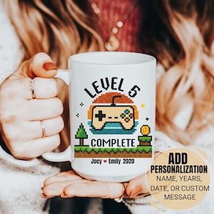 May include: White ceramic mug with a pixelated video game controller design. The mug features the text "LEVEL 5 COMPLETE" with a pixelated landscape and the names "Joey & Emily 2020". The mug also has a callout for personalization.