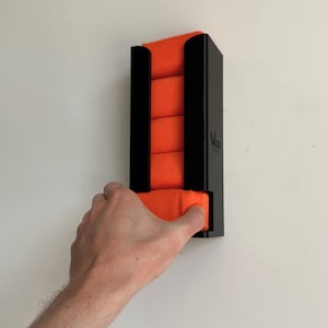 May include: A black wall-mounted organizer with orange fabric rolls. The organizer has a rectangular shape with vertical black supports. The brand name "Vast" is visible on the side. A hand is pulling an orange fabric roll from the bottom.