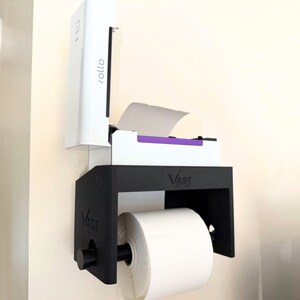 Op de afbeelding: Een aan de muur gemonteerde labelprinter in wit en zwart. De printer heeft het woord "rollo" op de zijkant gedrukt. Een grote rol wit papier wordt op zijn plaats gehouden door een zwarte beugel met het woord "VAST" erop.