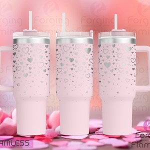 Puede incluir: Tres vasos de acero inoxidable de color rosa con un patrón de corazones plateados. Los vasos tienen una tapa y un asa. Los vasos están sobre un fondo rosa y blanco.