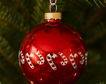 May include: A red Christmas ornament with a gold cap and a silver hook. The ornament is decorated with white candy cane designs. The ornament is hanging on a Christmas tree.