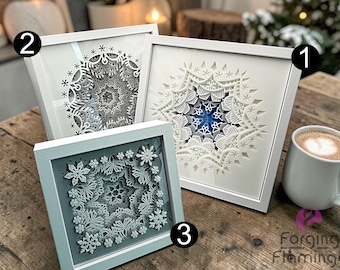 May include: Three framed paper cut art pieces with intricate snowflake designs. The frames are white and the artwork features layers of white paper with a blue accent in one. A white mug with a coffee drink is on the right.