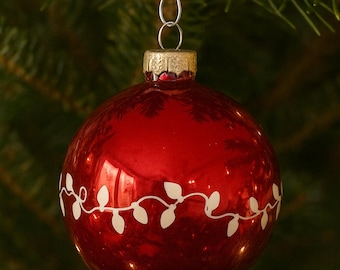 May include: A shiny red Christmas ornament with a white, stylized garland design. The ornament has a gold-colored cap and a silver hook for hanging. The ornament is hanging on a green Christmas tree.