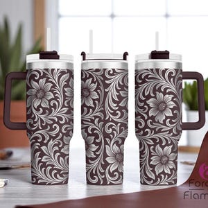 May include: Three stainless steel tumblers with floral patterns. The tumblers have a dark brown background with a white floral design. Each tumbler has a handle, a white lid, and a straw. The tumblers are on a light-colored surface.