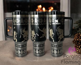 May include: Three black stainless steel travel mugs with a gold-colored design. The mugs feature a festive, wintery pattern with a repeating design of a stylized Christmas tree and a small snowflake. The mugs have a black handle and a black lid. The mug on the right has the word "STANLEY" printed on it.