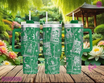 May include: Three green travel mugs with handles and straws, featuring a tarot card design in silver. The cards include images of frogs, stars, and moons. The mugs are set on a wooden surface with a blurred background of a garden.