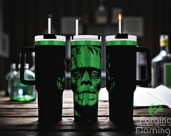 May include: Black insulated travel mugs with a green Frankenstein's monster graphic. The mugs have white lids, black handles, and a straw. One mug has a lit candle in the straw hole. The mugs are on a wooden table.