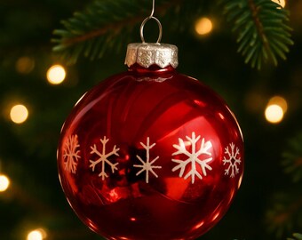 May include: A shiny red Christmas ornament with white snowflake designs hangs from a green pine branch. The ornament has a silver cap and is suspended by a wire. The background is blurred with warm, glowing lights.