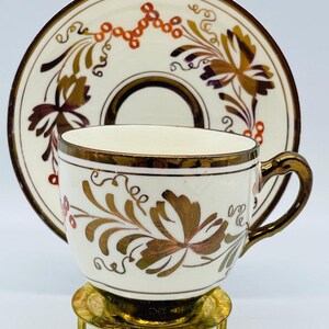 May include: A white teacup and saucer set with gold and brown floral designs. The cup and saucer have gold trim. The cup is on a gold-colored metal stand. The saucer is behind the cup.