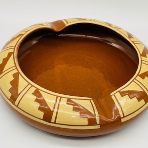 May include: A brown ceramic ashtray with a geometric design in a lighter shade of brown. The design features a series of triangles and squares.